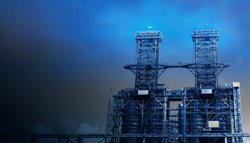 Industrial facility with two large metal structures, scaffolding, and pipes beneath a clear blue sky.
