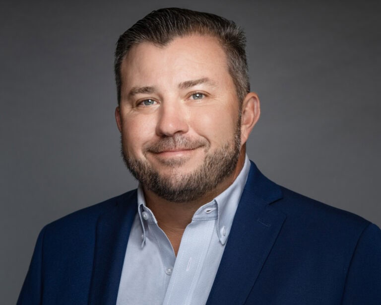 Man with short brown hair and beard in a blue suit jacket, smiling confidently against a gray background, showing leadership.